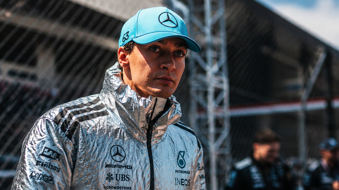 Piloto de Fórmula 1 com casaco prateado e boné azul da Mercedes num paddock de corrida.